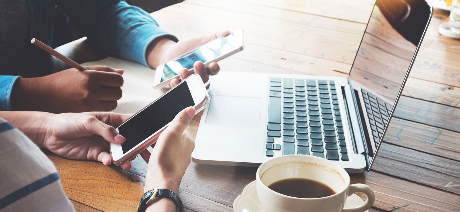Hands using a phone and laptop on a wooden table beside a coffee
