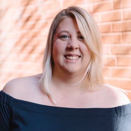 Blonde woman in an off-the-shoulder top smiling in front of a brick wall