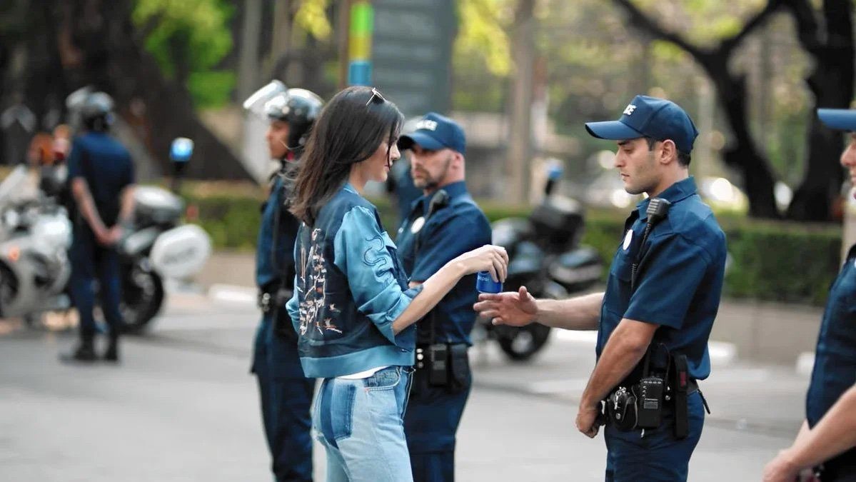 A woman offering a drink can to an officer in a line of riot police