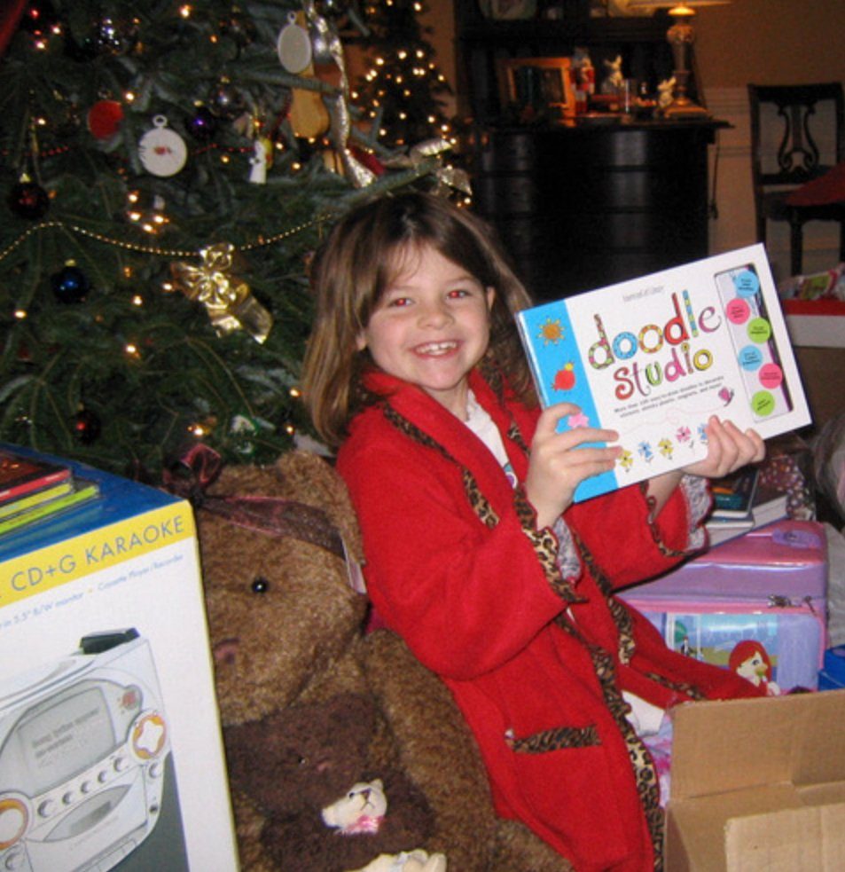 A girl in a red coat holding up a Doodle Studio art kit beside a Christmas tree