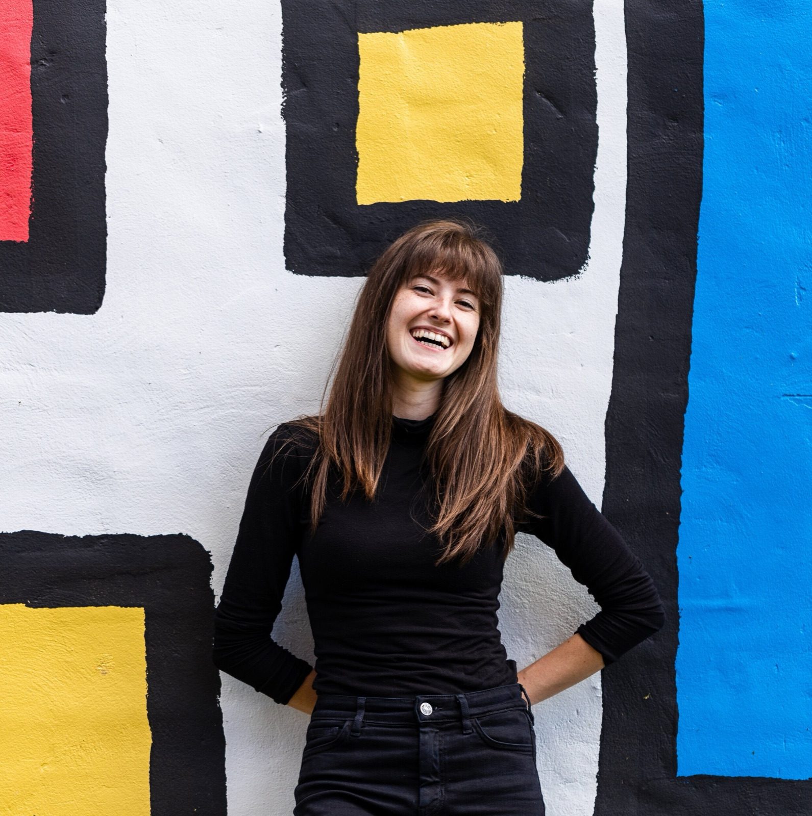 Morgen Tanksley in black smiling in front of a mural of yellow, red and blue squares
