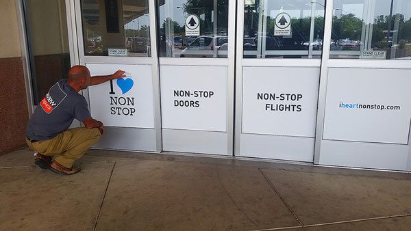 Installer applying I heart nonstop decals to the airport's glass doors, labeled Non-Stop Doors and Non-Stop Flights