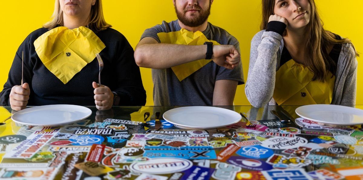 Three people in yellow bibs holding cutlery at a table covered in stickers