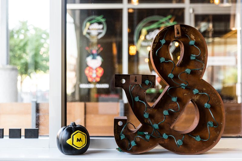 Rusted metal ampersand strung with lights beside a black Maycreate pumpkin