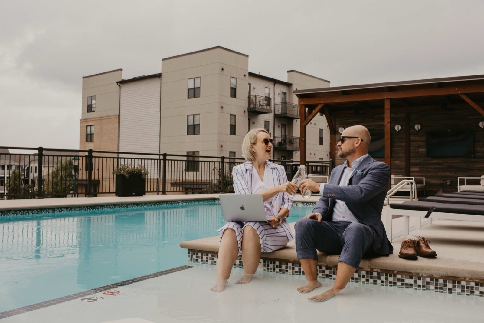 Two people sitting at the edge of a swimming pool with a laptop, an apartment block behind them