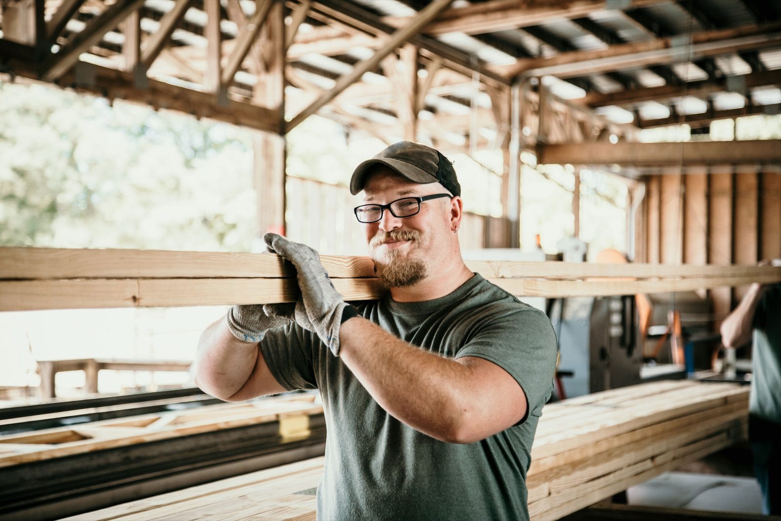 A worker carrying a length of timber on his shoulder in an open-sided lumber shed
