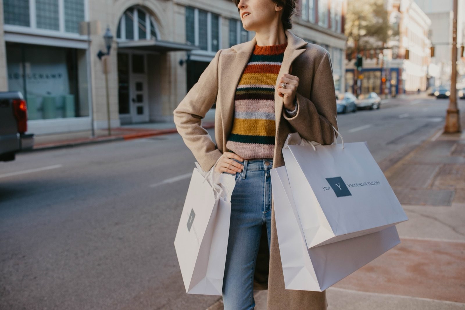A woman carrying shopping bags across a downtown street in a striped jumper and camel coat