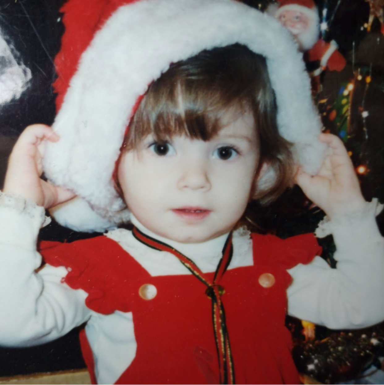 A toddler in red overalls pulling a Santa hat down over her head