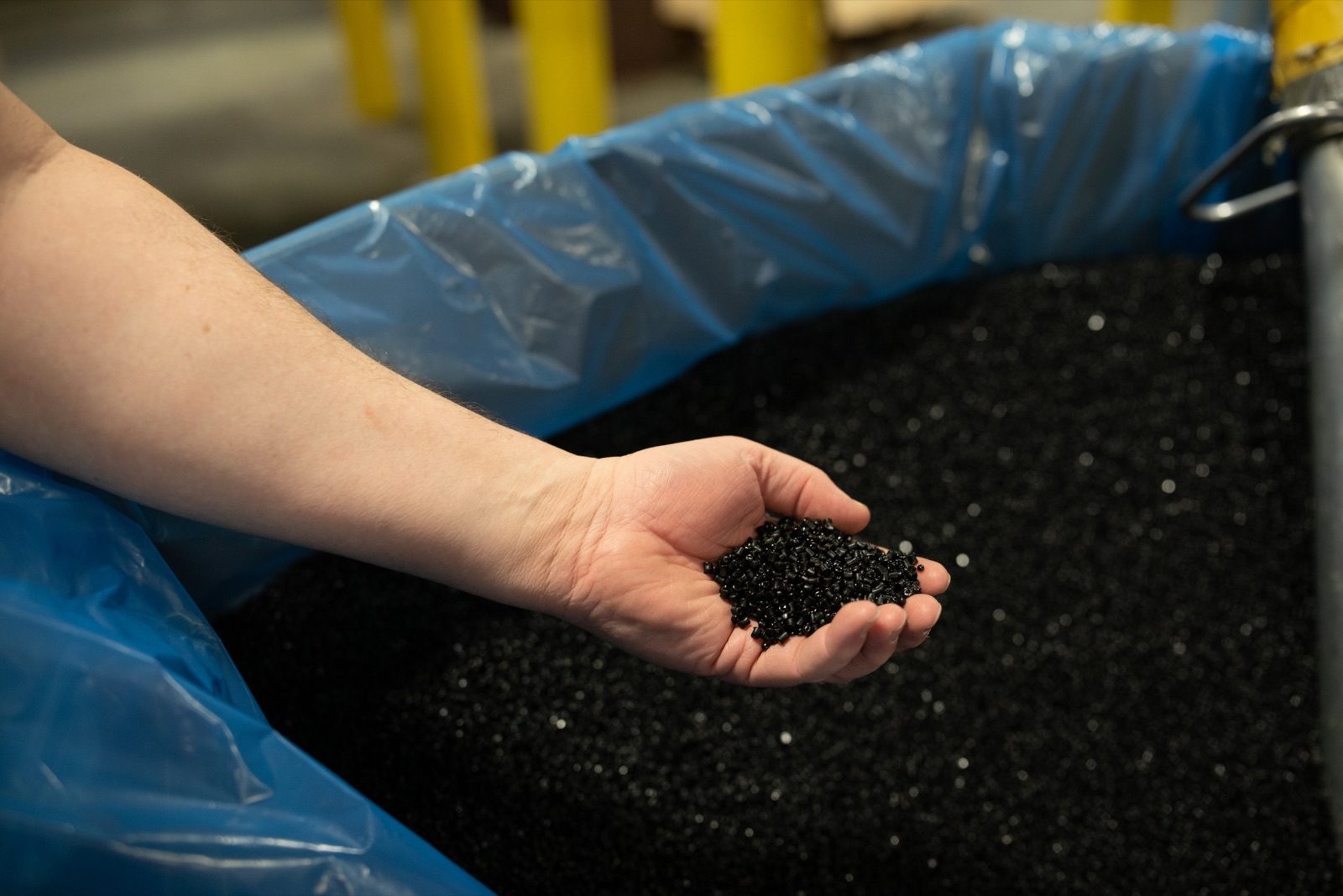Hand holding a scoop of black plastic pellets from a lined bin