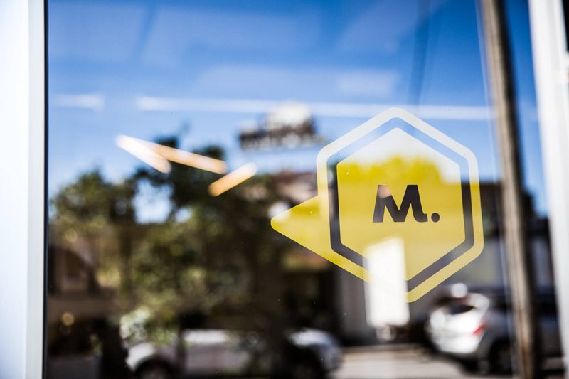 Maycreate M logo decal on a window reflecting the street