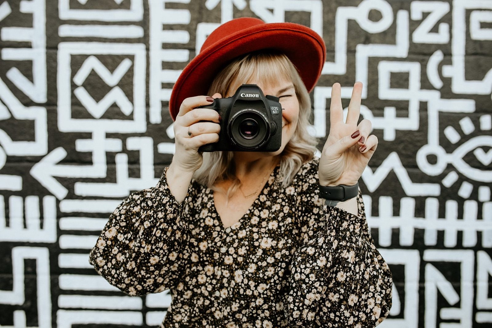 Lindsey Lowe in a red hat holding a camera and flashing a peace sign in front of a black and white mural
