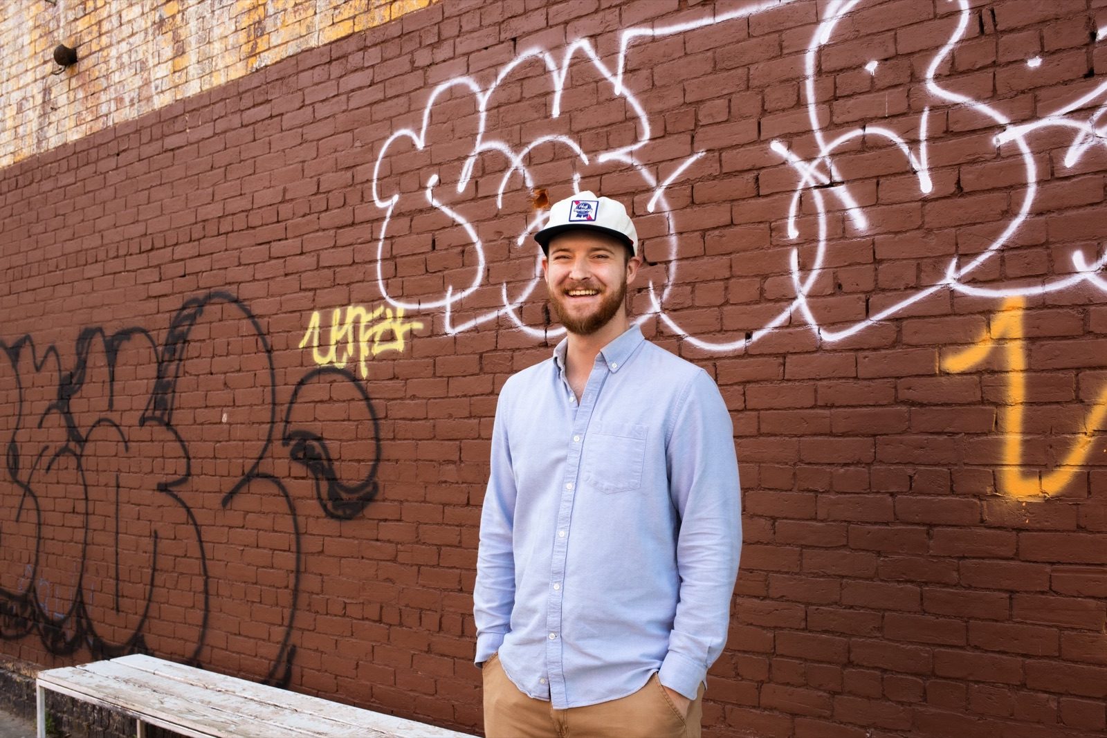 Ian Lewis in a light blue shirt and white cap in front of a graffiti-covered brick wall