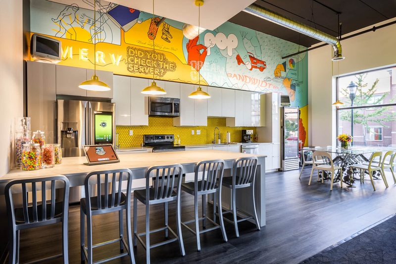 Office kitchen with a yellow tile backsplash, a mural and a long counter with metal stools