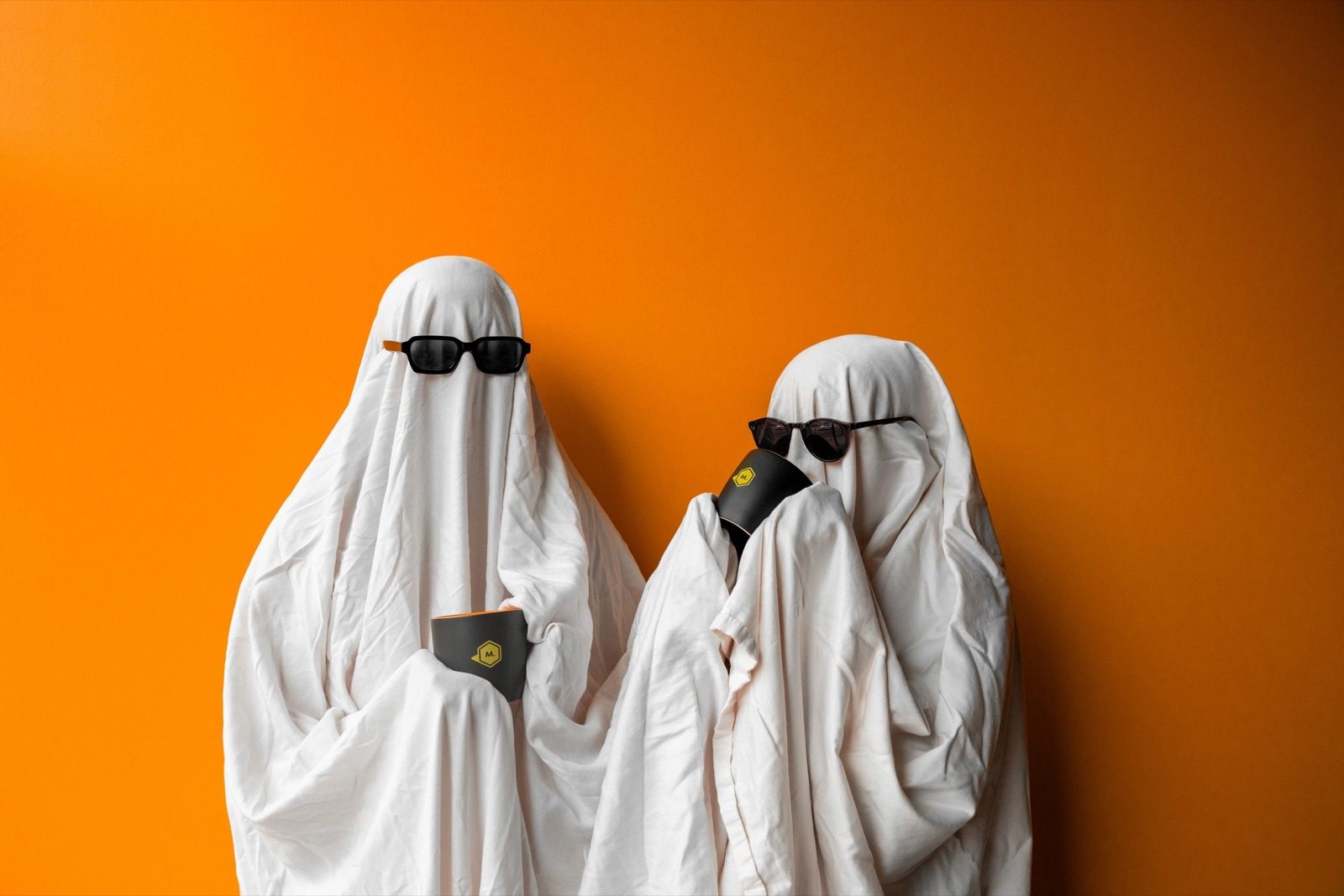 Two people in white sheet ghost costumes and sunglasses against an orange background