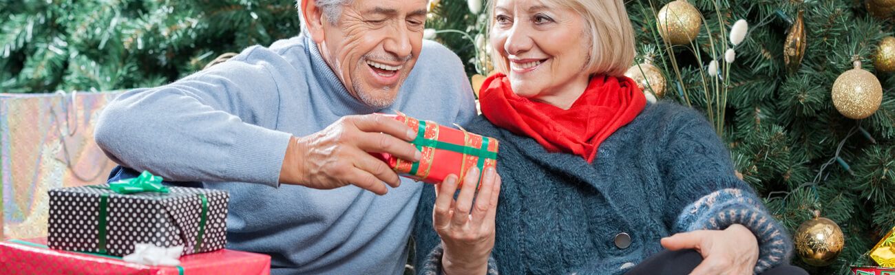 Older couple exchanging a wrapped gift beside a Christmas tree