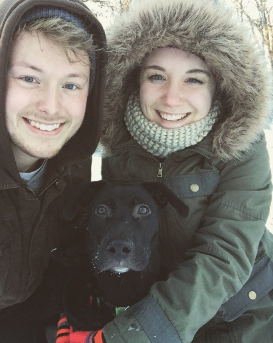 A couple in heavy winter coats crouched in the snow with a black labrador
