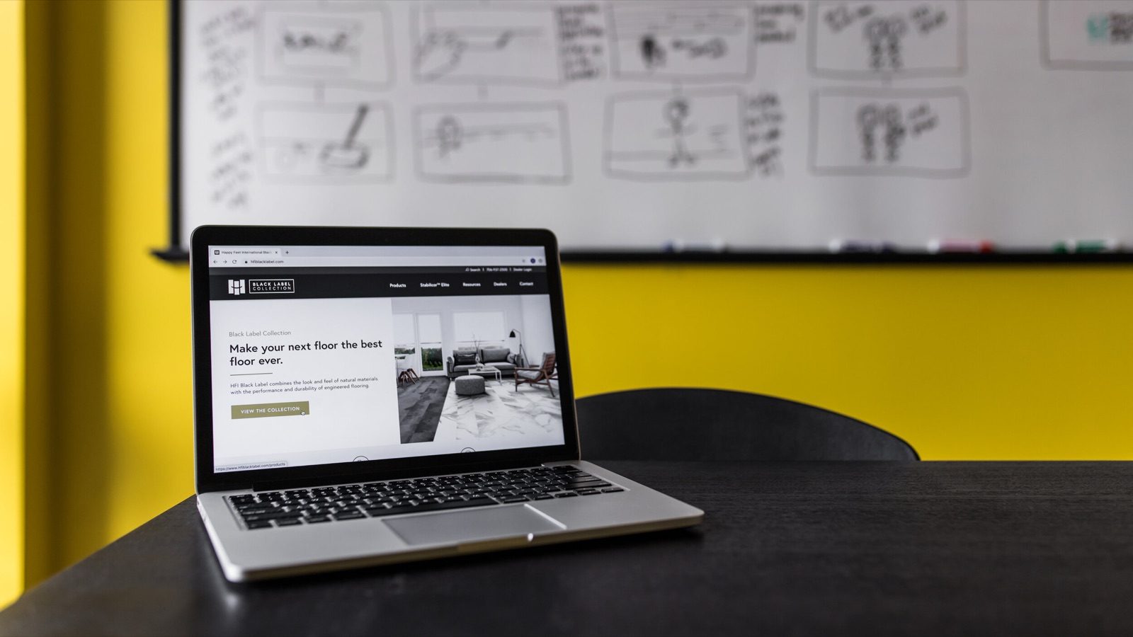 Laptop on a desk showing a website homepage, storyboard sketches pinned on the wall behind