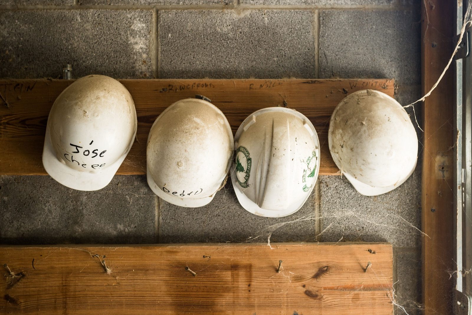 Four well-worn hard hats hanging on a wooden rail, each marked with a name