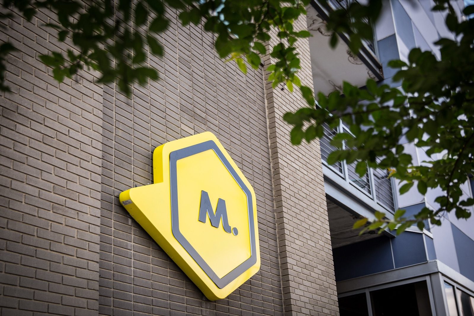 Yellow Maycreate M sign on a brick wall beside leafy branches