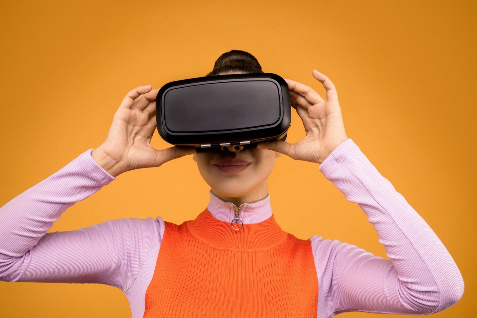 Woman in a lilac top wearing a virtual reality headset against an orange background