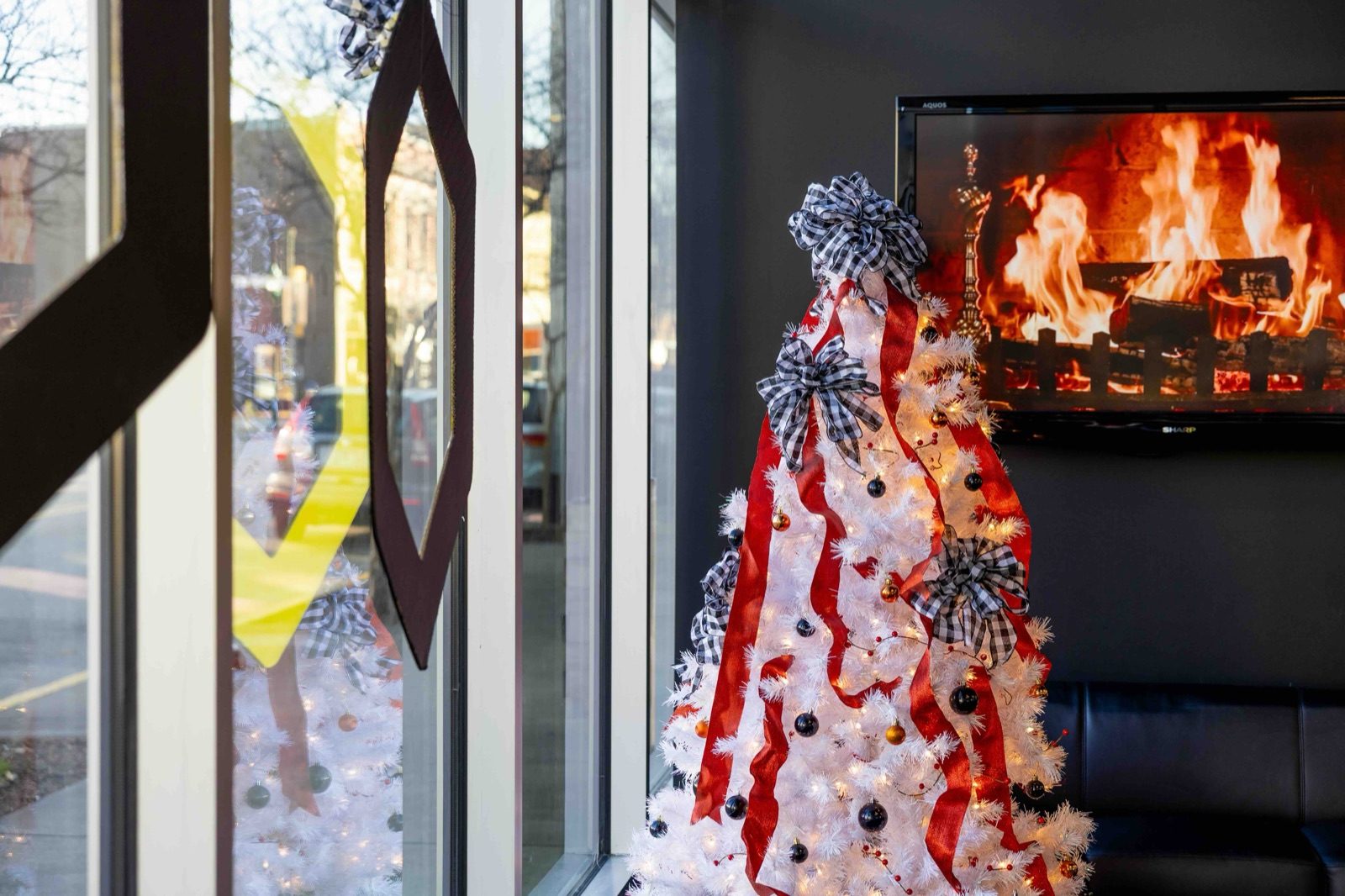 A white Christmas tree trimmed with red ribbon beside an office window, a fireplace playing on a screen behind it