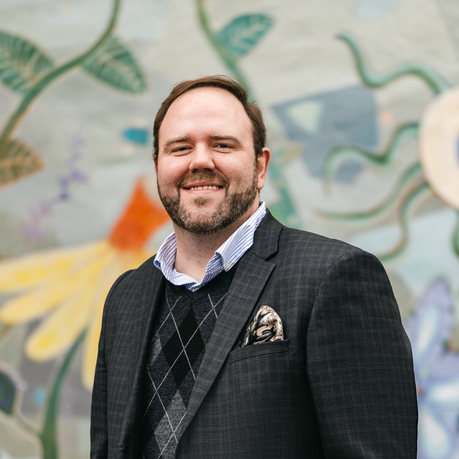 Man in a checked blazer and sweater vest smiling in front of a floral mural