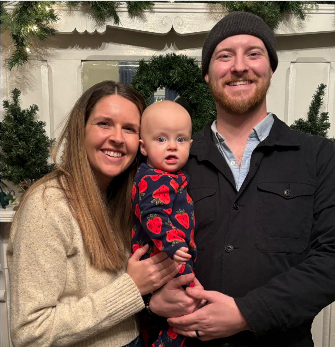 A couple holding a baby in patterned pyjamas in front of a mirror hung with evergreen garland