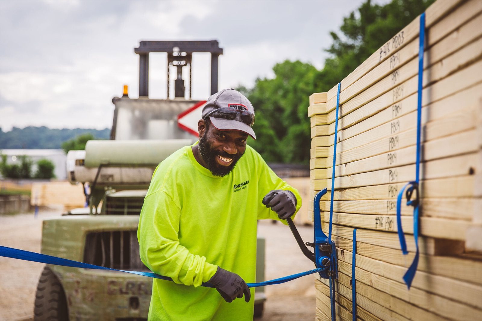 A worker strapping a bundle of timber on a truck bed with a forklift behind