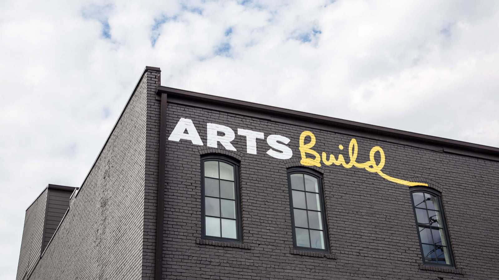 ArtsBuild sign in white block letters and yellow script on a dark brick building