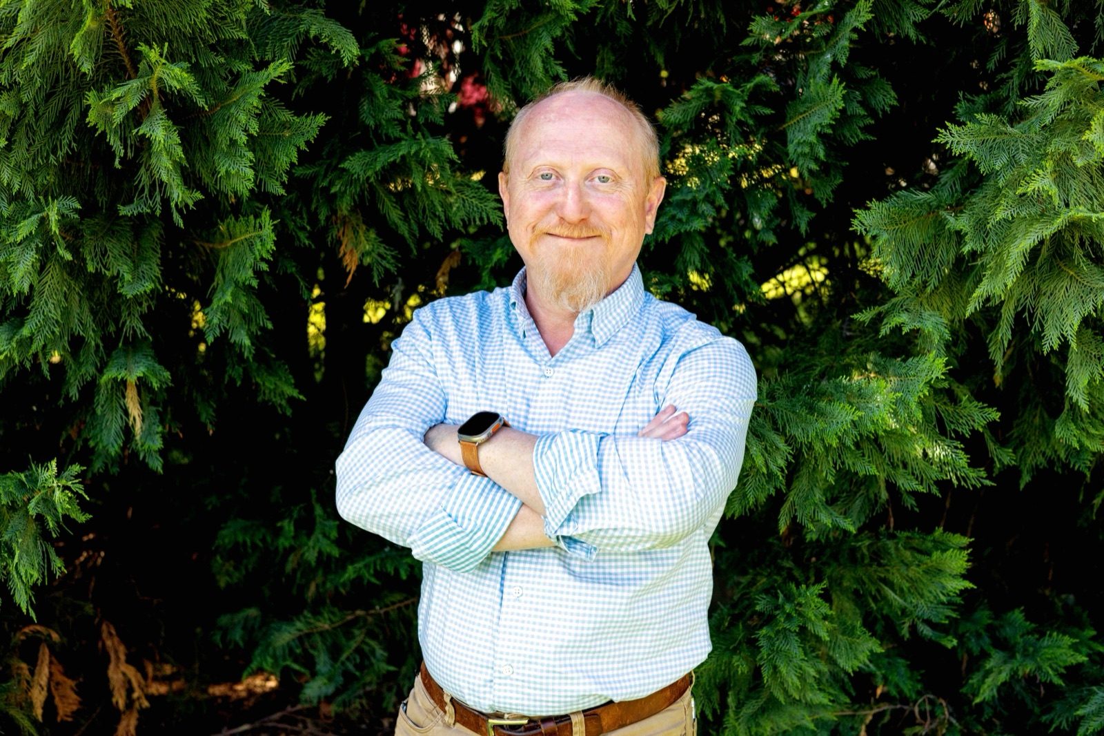Glen Austin standing with his arms folded in front of an evergreen hedge