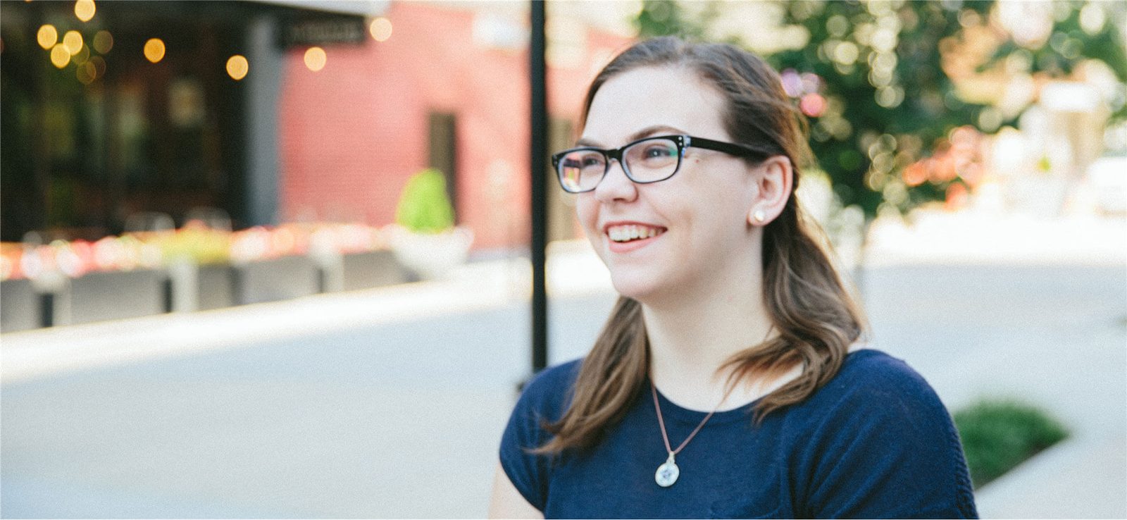 Smiling woman in glasses and a navy top outdoors with string lights behind