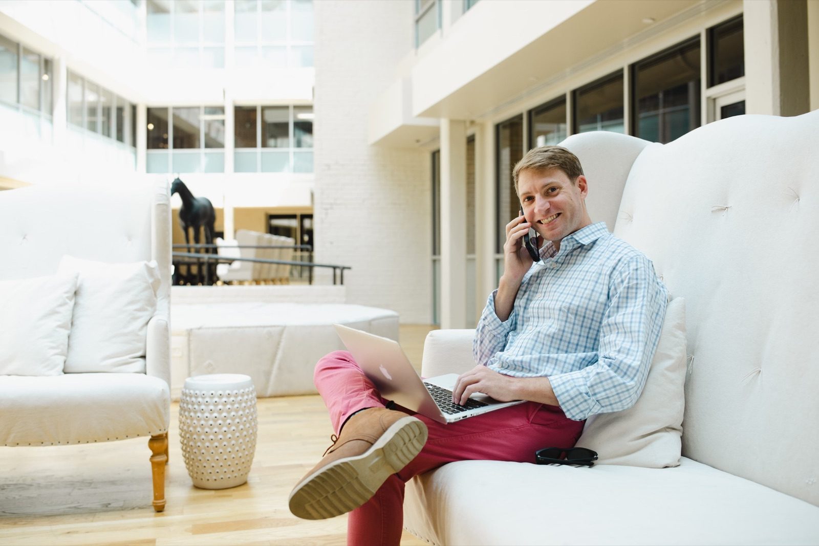 Beau on a white sofa in a bright lobby, phone to his ear and laptop on his knee