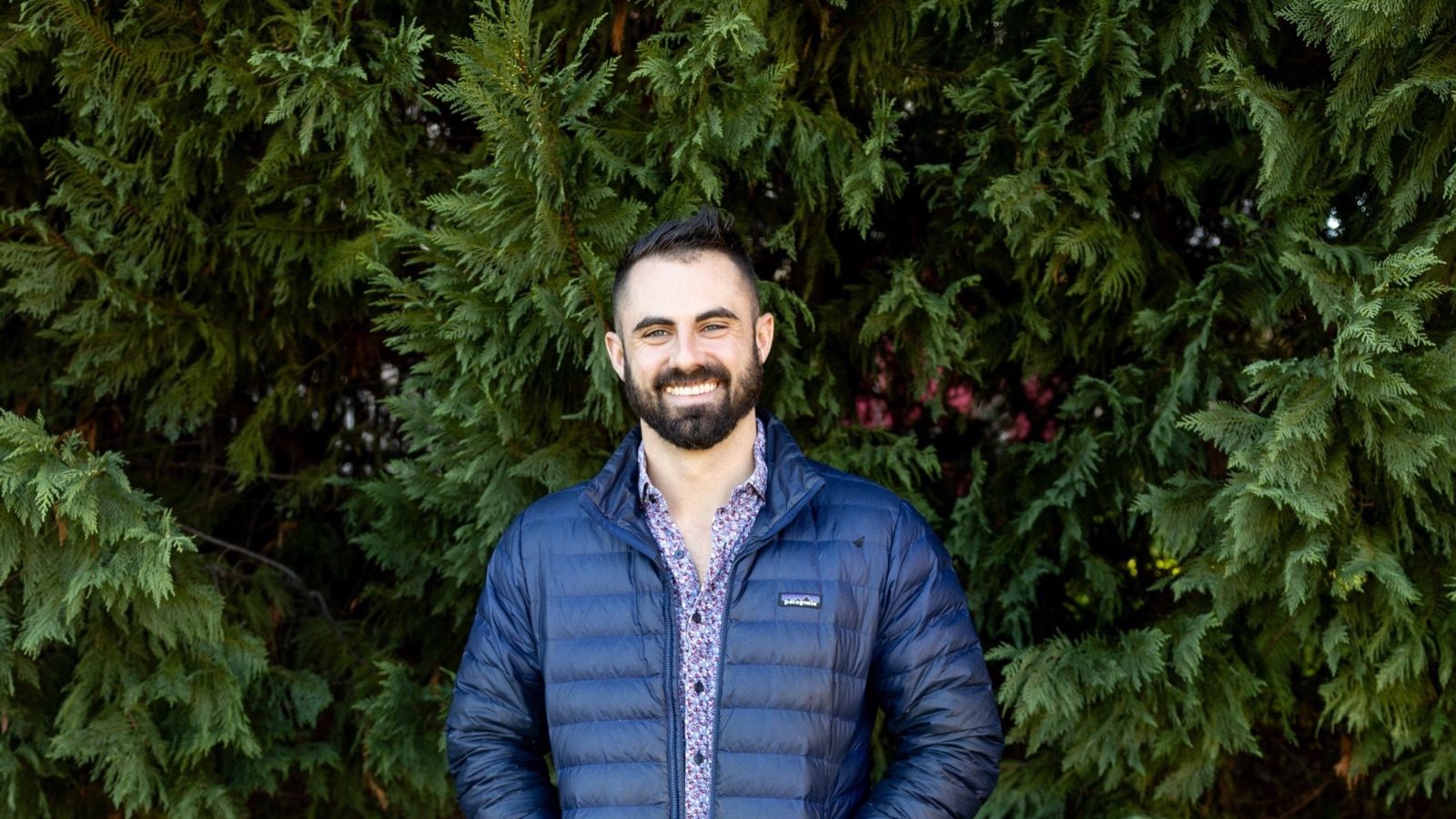 Chris Matthews smiling in front of an evergreen hedge