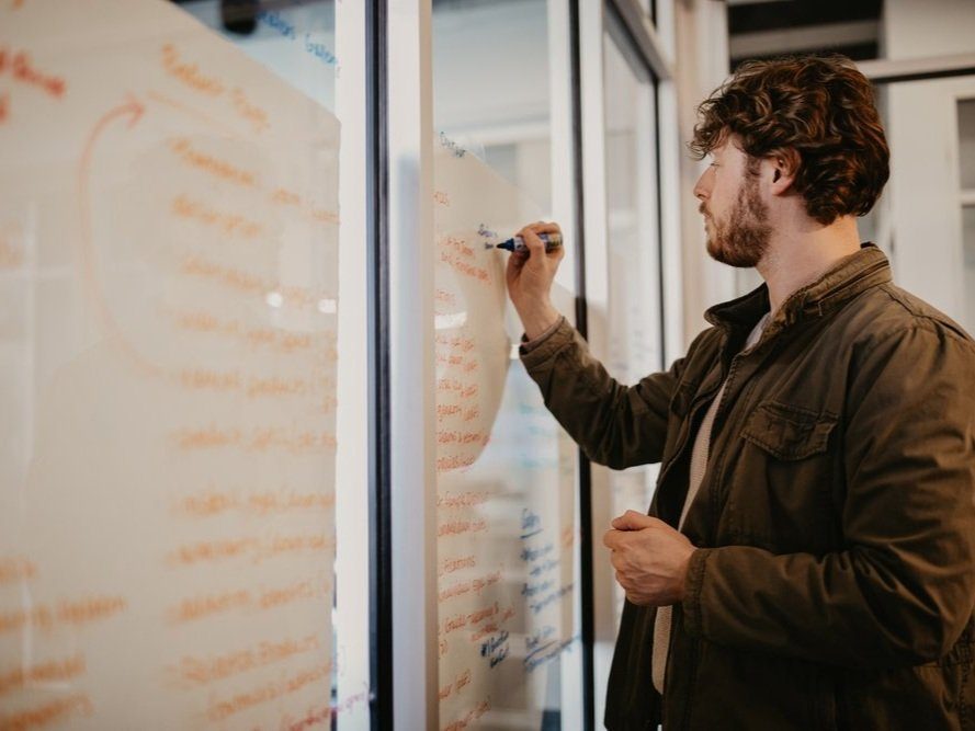 Man in a brown shirt writing on a glass wall covered in notes