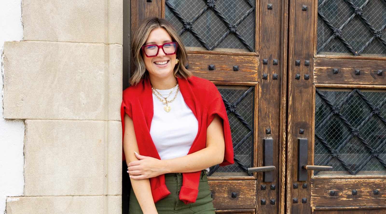 Lilee Rose in a red cardigan and sunglasses, leaning against a pair of carved wooden doors