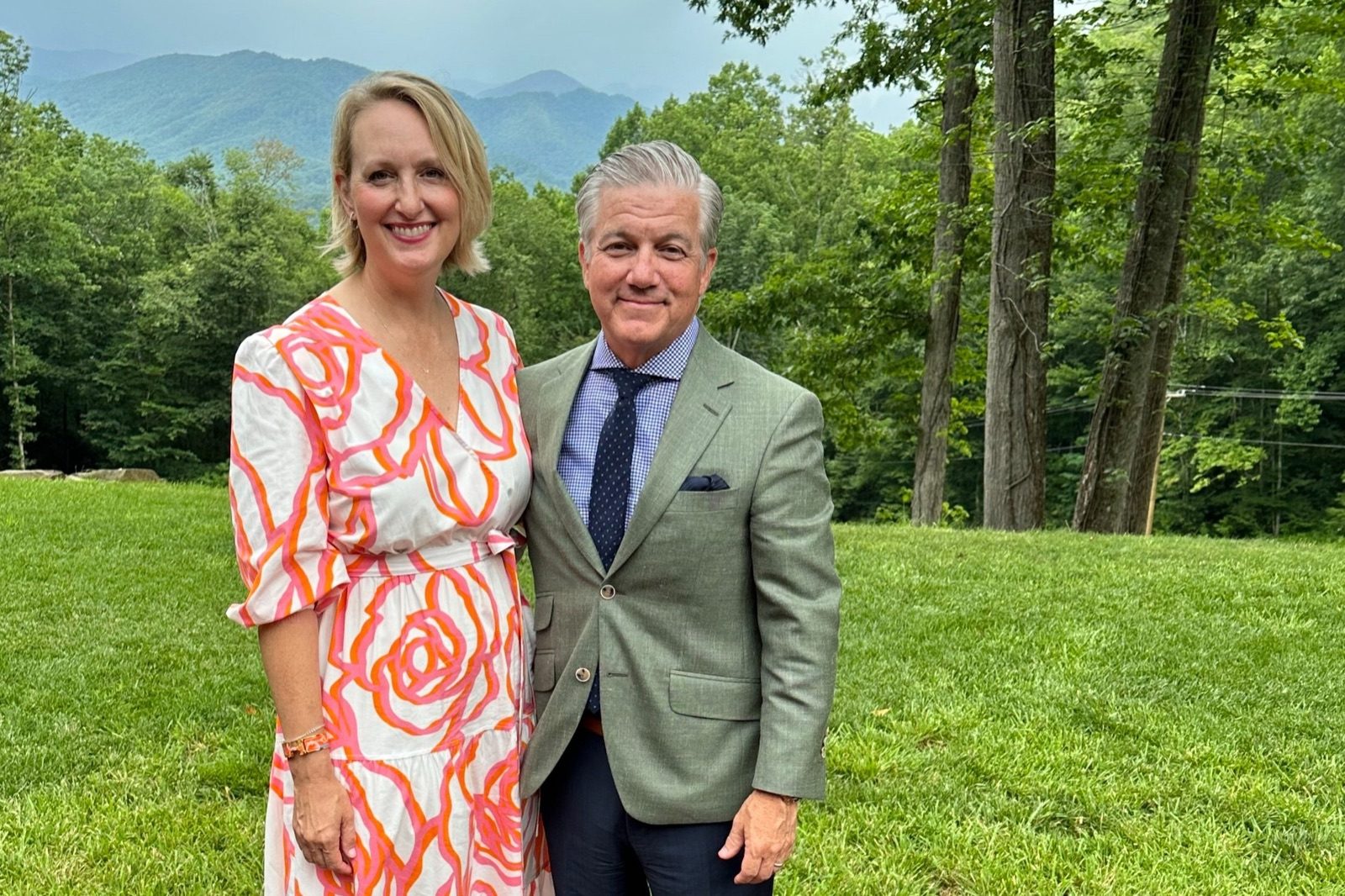 Woman in an orange floral dress and man in a green blazer standing on a lawn with mountains behind