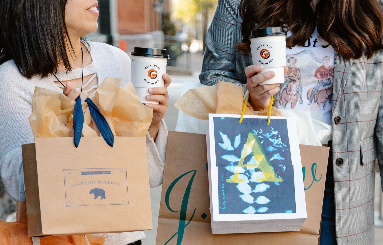 Two shoppers holding coffee cups and paper gift bags on a city sidewalk