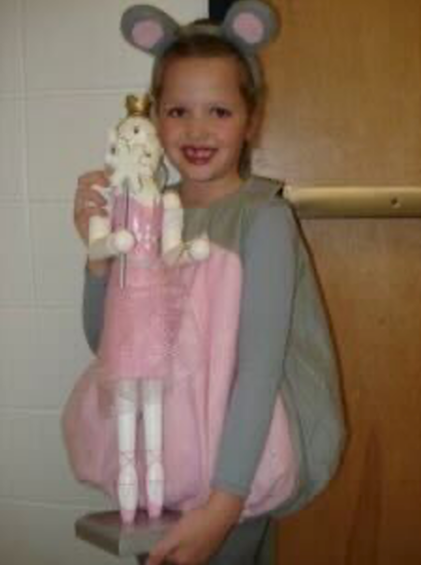 A child in a grey mouse costume and pink tutu holding a ballerina doll